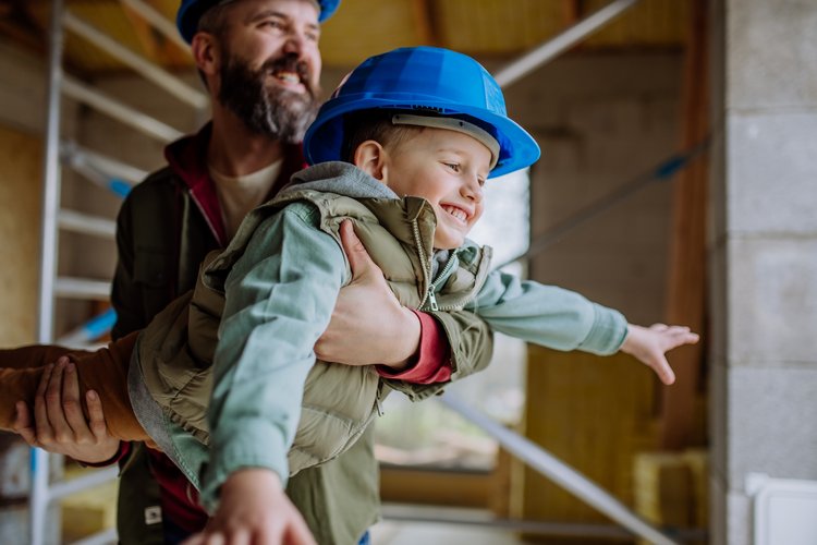 Ein Mann hält ein lachendes Kind mit blauem Bauhelm auf einer Baustelle im Rohbau.