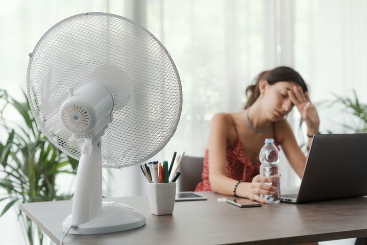 Eine Frau sitzt an einem Schreibtisch in einem aufgeheizten Raum und wirkt erschöpft. Vor ihr steht ein Tischventilator, der für etwas Luftbewegung sorgt. Sie hält eine Wasserflasche in der Hand und stützt die Stirn, während sie vor einem Laptop arbeitet. Die Szene vermittelt starke Sommerhitze im Innenraum und Belastung durch hohe Temperaturen.