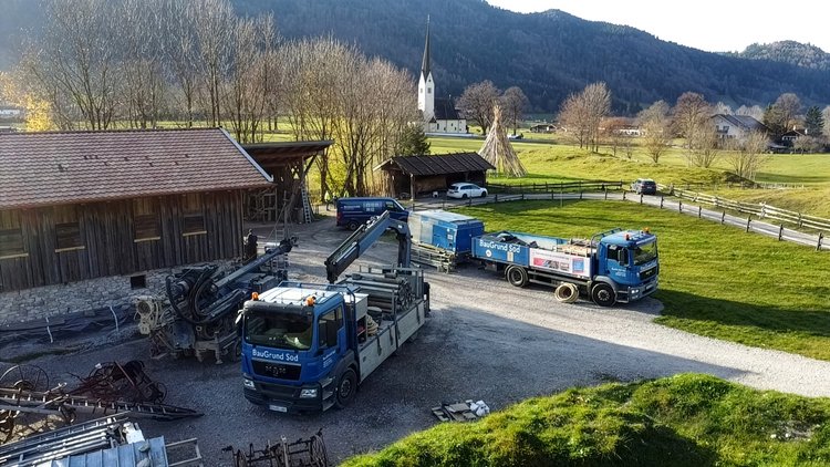Ländliche Baustellenszene mit Bohrgerät und Firmenfahrzeugen vor einem Holzgebäude, umgeben von Wiesen; im Hintergrund eine Kirche und bewaldete Hügel.