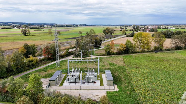 Luftaufnahme eines Umspannwerks in ländlicher Umgebung mit Feldern, Straßen und Stromleitungen unter bewölktem Himmel