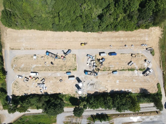 „Drohnenaufnahme einer Baustelle mit mehreren Baumaschinen, Fahrzeugen und Materiallagern auf einer länglichen, unbefestigten Fläche, umgeben von dichtem Grün und einer Straße im Vordergrund.“