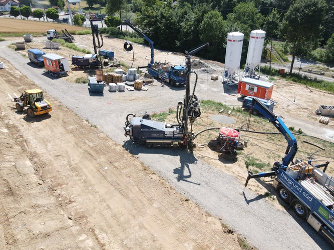 Baustelle aus der Vogelperspektive mit mehreren Bohrgeräten, Kränen und Arbeitsfahrzeugen, Materialstapeln sowie Silos auf einer geschotterten Fläche, umgeben von Bäumen und Feldern.
