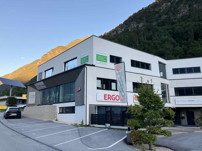 Modernes Geschäftsgebäude mit weißer Fassade, großen Fenstern und sichtbaren Firmenschildern wie ERGO und MEGA Textil, vor Berglandschaft mit Parkplatz im Vordergrund.