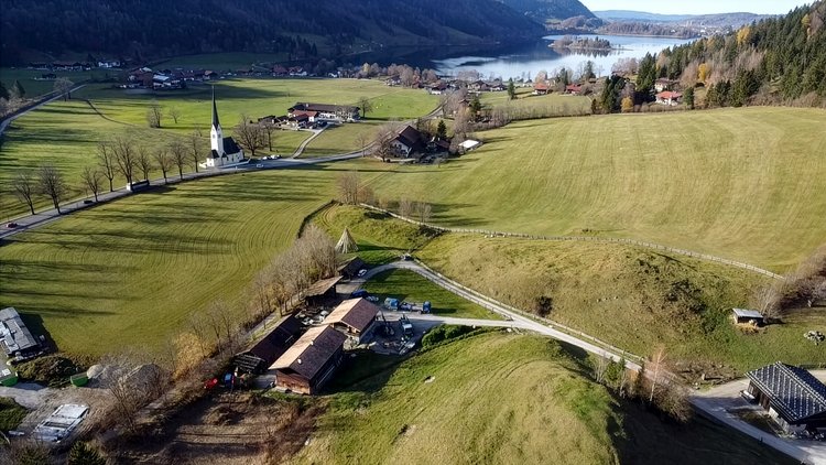 Luftaufnahme einer ländlichen Landschaft mit Wiesen, Bauernhöfen und einer Kirche, im Hintergrund ein See und bewaldete Hügel.