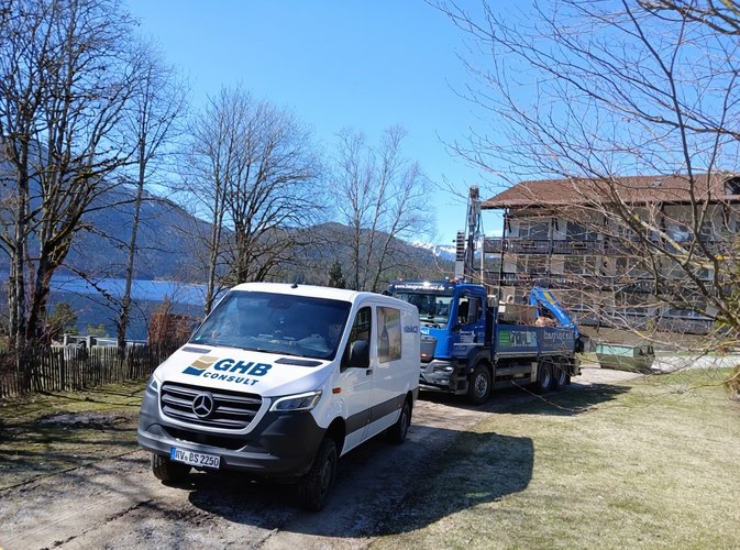 Firmenfahrzeuge von GHB Consult auf einem Grundstück am Bodensee: ein weißer Transporter und ein Lkw mit Bohrgerät vor einem Gebäude, umgeben von Bäumen und Berglandschaft.